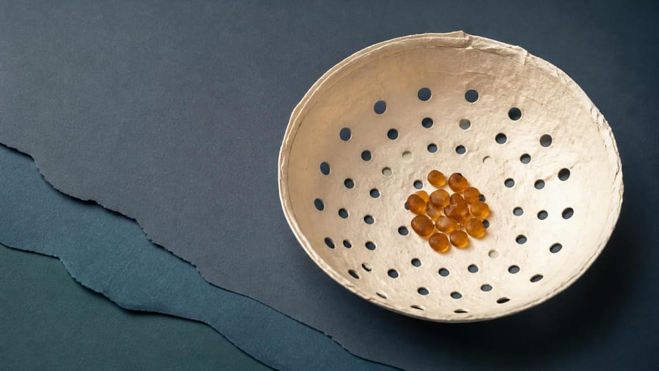 Perforated cut-paper bowl retaining a small cluster of amber pebbles.